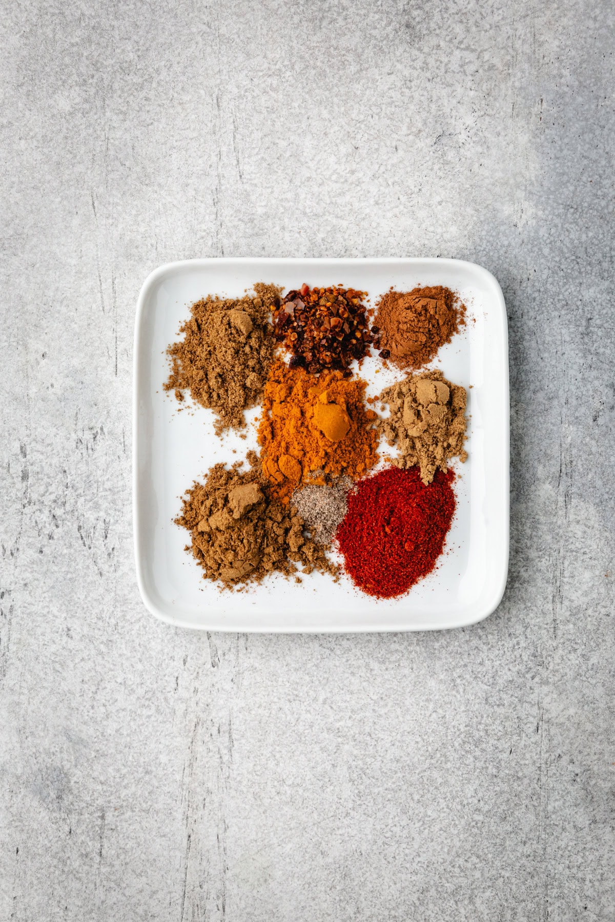 A white square plate holds eight piles of shawarma spices varying in color and texture, arranged neatly against a textured gray background.