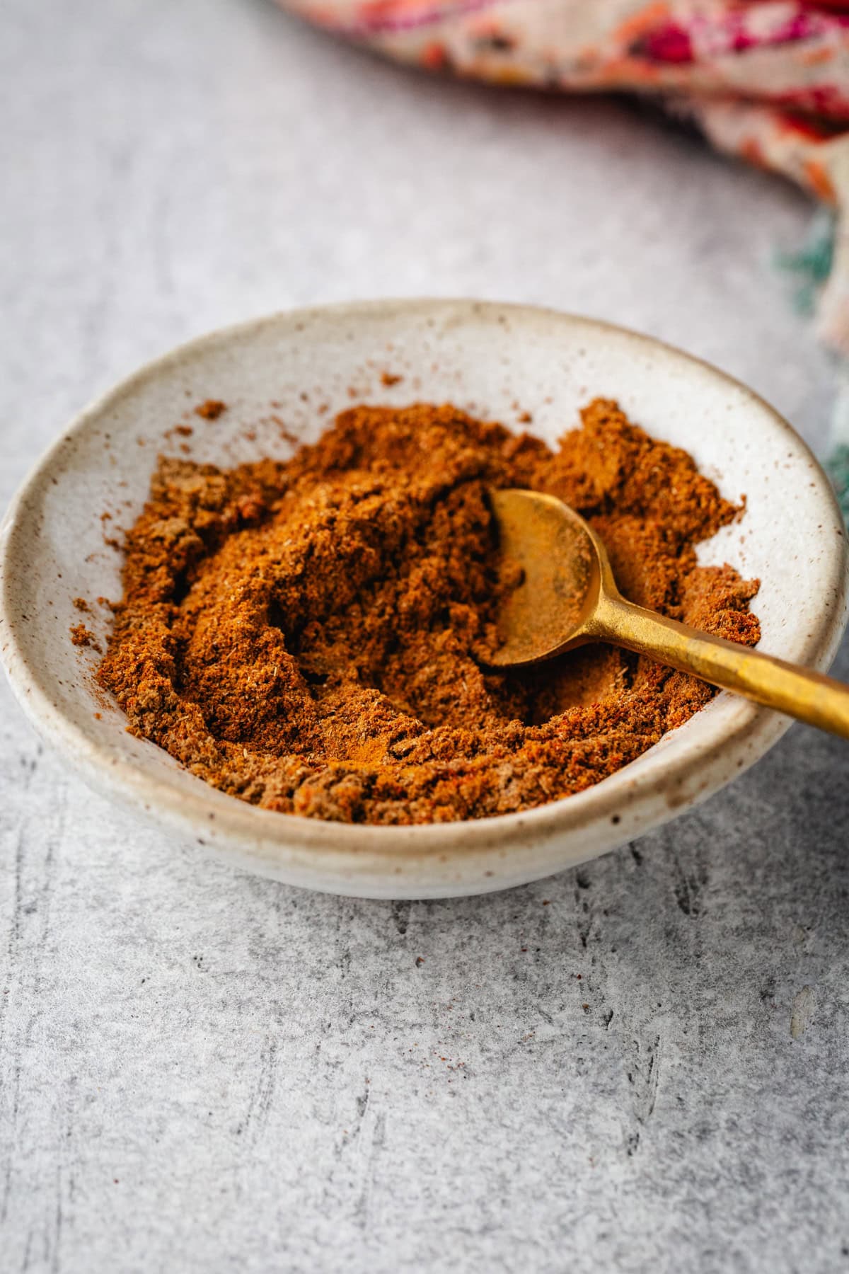 A small white bowl filled with a pile of shawarma spice mix, accompanied by a brass spoon, sits on a gray textured surface. A hint of a colorful cloth is visible in the background.