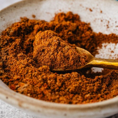 A bowl filled with ground spices is shown with a gold spoon resting in it. The spices are a rich brown color, and the bowl is placed on a speckled gray surface.