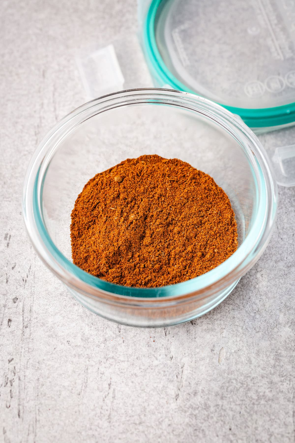 A glass bowl filled with shawarma spice mix is placed on a textured gray surface. The bowl has a light green lid that is partially open. The cinnamon is finely ground and evenly distributed inside the bowl.
