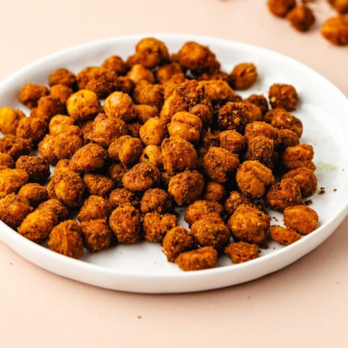A white plate filled with crispy, turmeric roasted chickpeas sits on a light beige surface. A few chickpeas are scattered around the plate, adding a touch of warmth and spice to the scene.