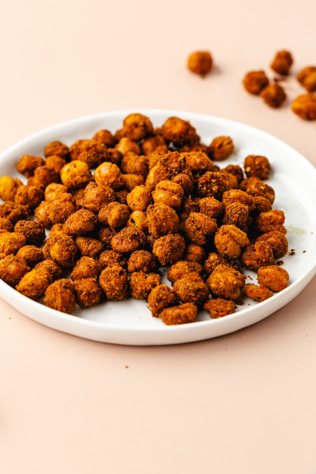 A white plate filled with crispy, turmeric roasted chickpeas sits on a light beige surface. A few chickpeas are scattered around the plate, adding a touch of warmth and spice to the scene.