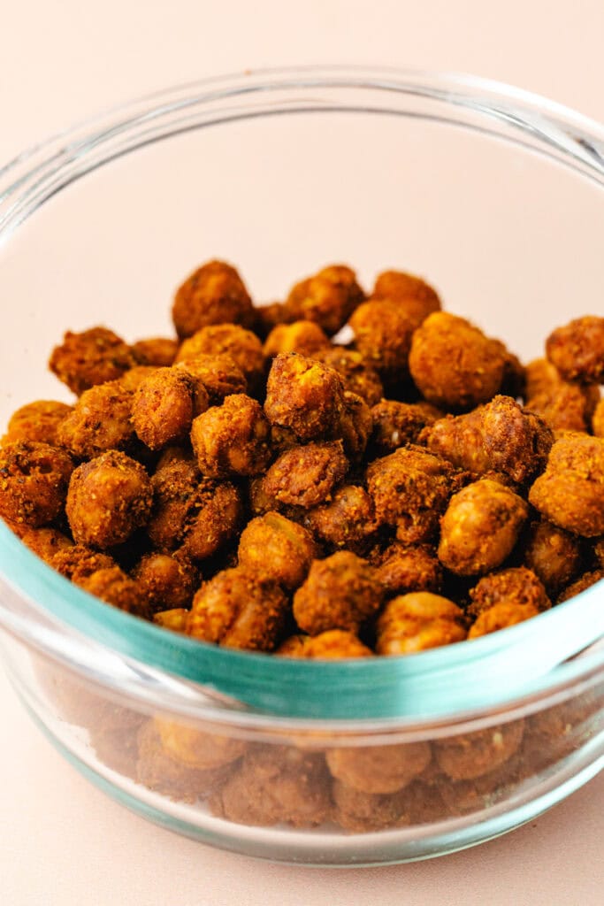 A glass bowl filled with turmeric roasted chickpeas, seasoned with spices, creating a golden-brown crispy texture. The chickpeas are piled up in the bowl, showcasing their crunchy and flavorful appearance.