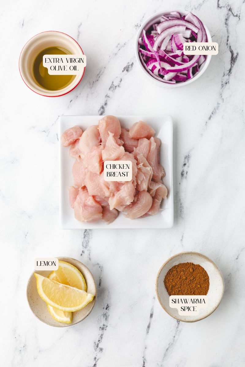 Top-down view of a chicken shawarma bowl being prepared on a marble surface: a square plate with raw chicken breast pieces, bowls with sliced red onion, extra virgin olive oil, lemon wedges, and a fragrant bowl of shawarma spice mix.