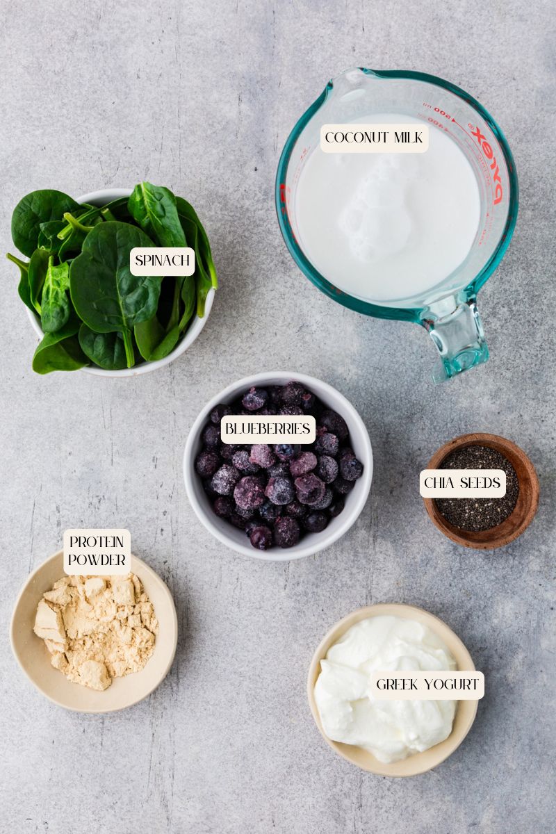 Top-down view of blueberry smoothie ingredients on a gray surface, featuring a measuring cup of coconut milk, a bowl of spinach, frozen blueberries, chia seeds, protein powder, and Greek yogurt.