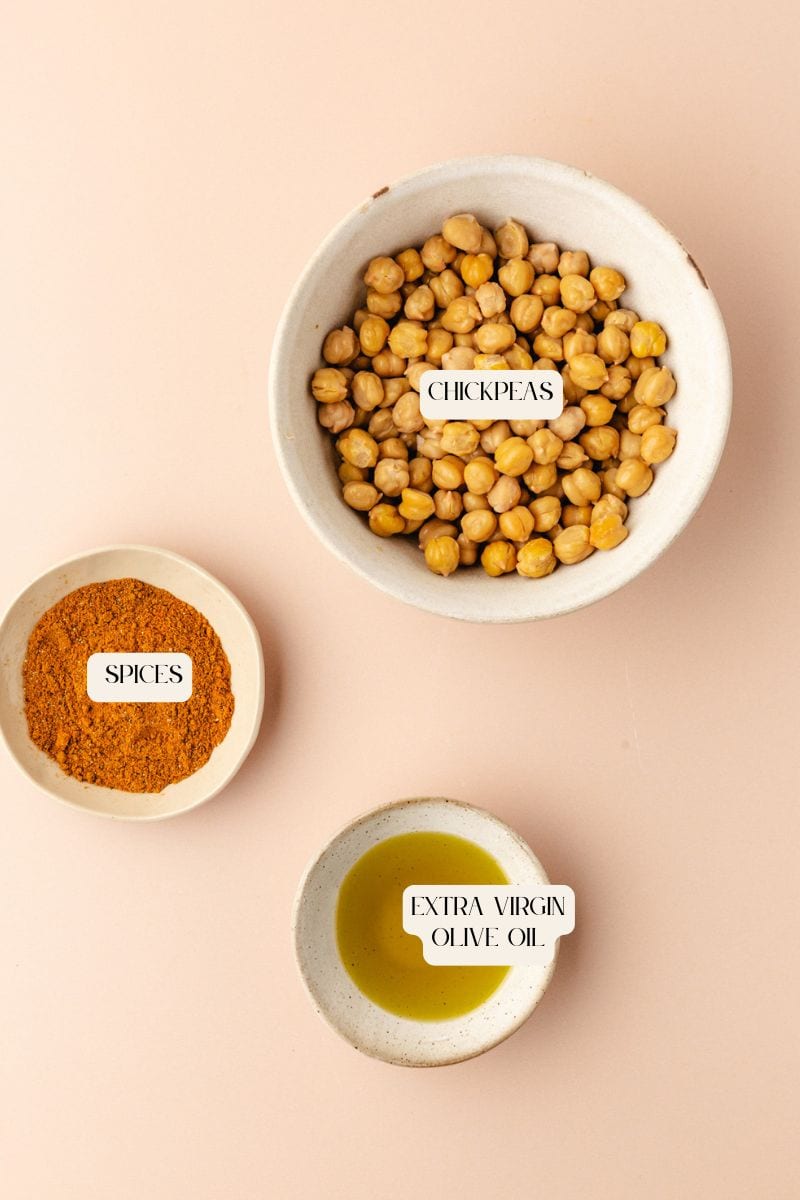 Three bowls on a light beige background: one with turmeric roasted chickpeas, another with ground spices, and the last with extra virgin olive oil. Each bowl is labeled accordingly.
