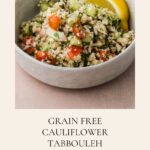 A bowl of grain-free Cauliflower Tabbouleh with chopped vegetables and parsley, garnished with two lemon wedges, sits on a table. The text below describes the dish as gluten free.