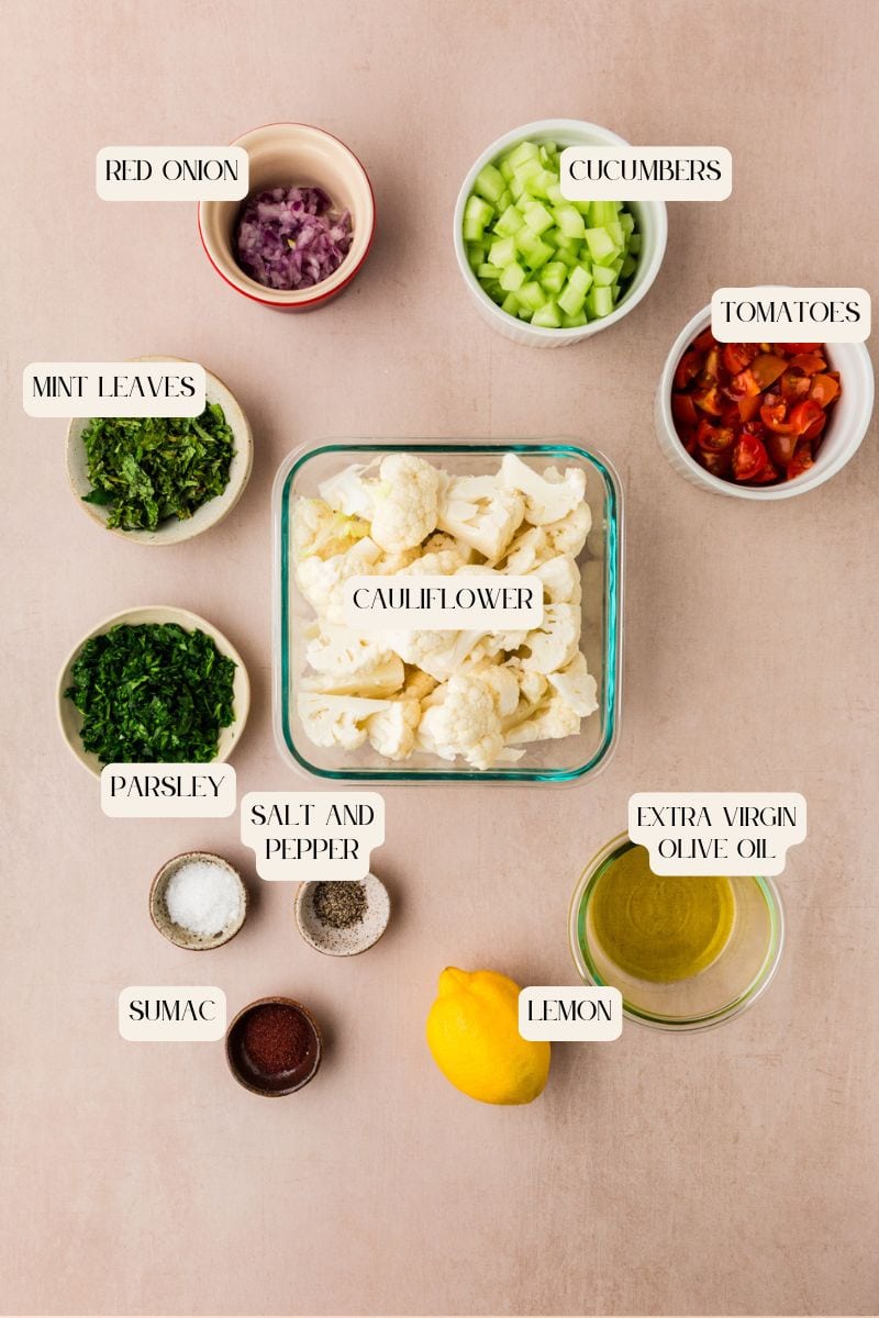 Tabbouleh ingredients are arranged on a beige surface: chopped red onion, cucumbers, tomatoes, mint leaves, parsley, and cauliflower florets. Accompanied by a lemon, extra virgin olive oil, salt, pepper, and sumac—all neatly divided in separate bowls.