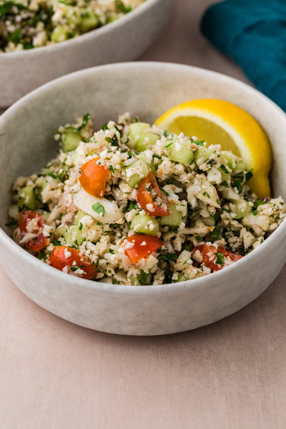 A bowl of tabbouleh salad with chopped cauliflower, cherry tomatoes, cucumber, and herbs. A slice of lemon is placed on the rim of the bowl. The dish is set on a light-colored surface.