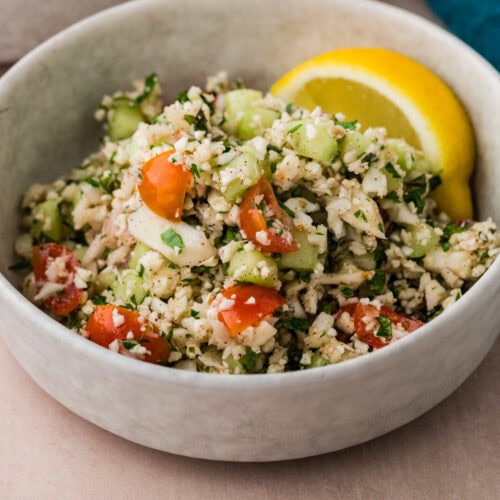 A bowl of fresh cauliflower tabbouleh salad features diced cucumbers, cherry tomatoes, and herbs. A lemon wedge adds a pop of color to this vibrant dish, set on a neutral-toned surface with just a hint of blue cloth in the background.