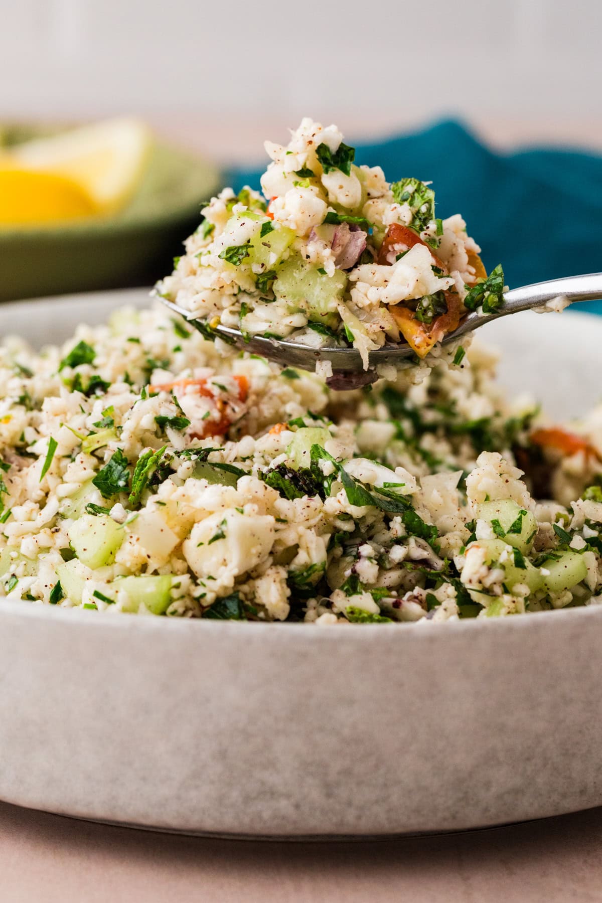 A close-up of a bowl filled with vibrant tabbouleh salad. This delightful mix features chopped cauliflower, cucumbers, parsley, and diced vegetables. A spoon lifts a portion above the bowl while a slice of lemon adds a zesty background touch.