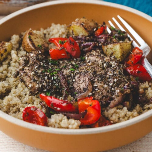 A bowl of quinoa topped with seasoned chicken pieces, roasted red peppers, and potatoes. The dish is garnished with herbs and served with a fork on the side.
