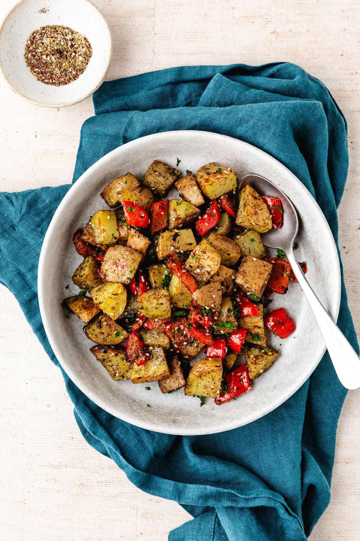 A bowl of zaatar potatoes and roasted red bell peppers rests on a gray plate, atop a teal cloth. A spoon lounges on the dish, while a small spice bowl peeks from the upper left corner. The inviting scene unfolds on a beige surface.