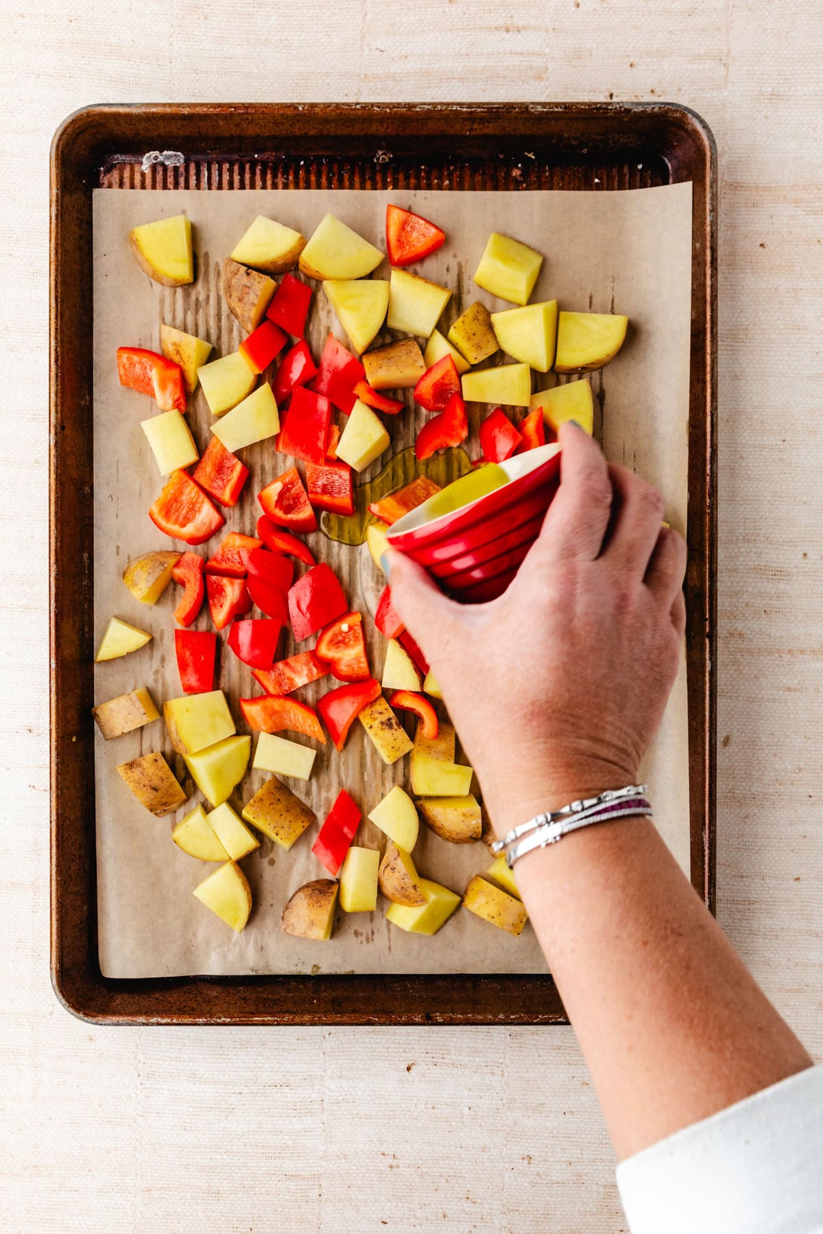 A hand is sprinkling zaatar seasonings from a red bowl onto a baking sheet filled with chopped potatoes and red bell peppers, placed on parchment paper. The enticing sheet rests on a beige surface.