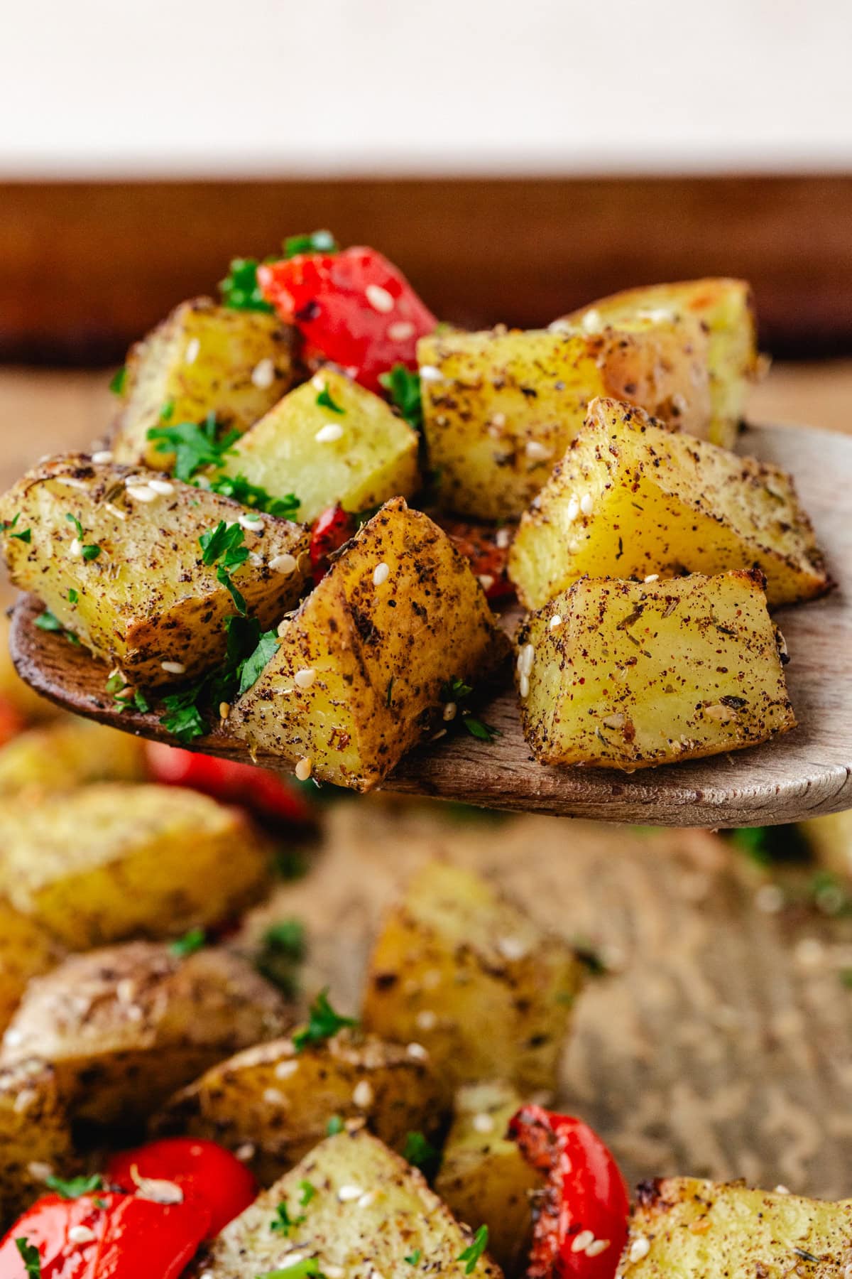Close-up of zaatar potato cubes garnished with fresh parsley and sesame seeds. Some pieces are skewered on a wooden fork, with others resting on a rustic wooden surface. Red pepper slices add a splash of color in the background.