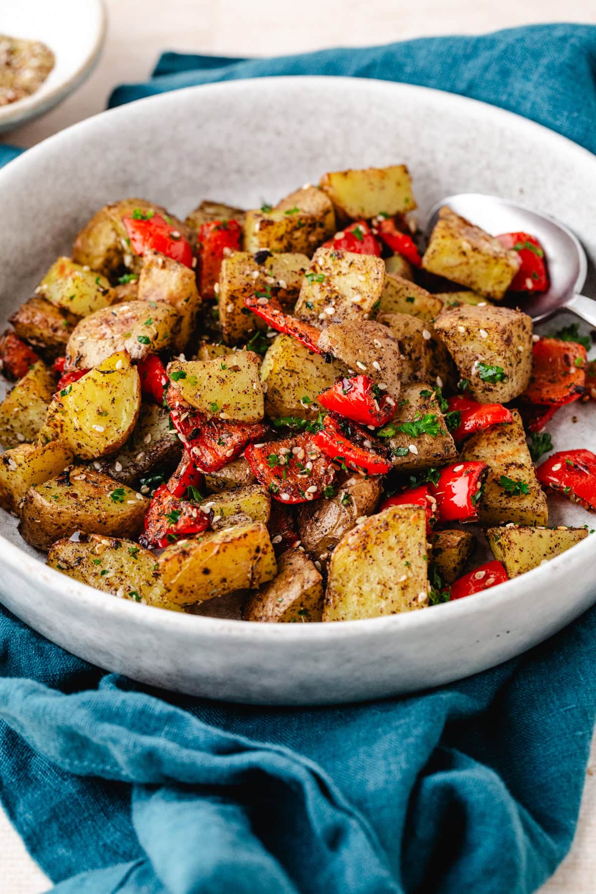 A bowl of zaatar potatoes and roasted red bell peppers, garnished with herbs and spices, sits on a blue cloth. A serving spoon rests in the dish, offering a vibrant and appetizing creation bursting with flavor.