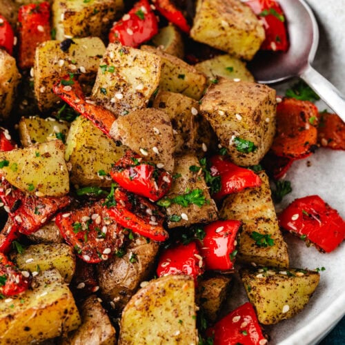 A close-up of a dish featuring zaatar potatoes with roasted red bell peppers, garnished with herbs and sesame seeds. The meal is served in a white bowl with a metal spoon on the side.
