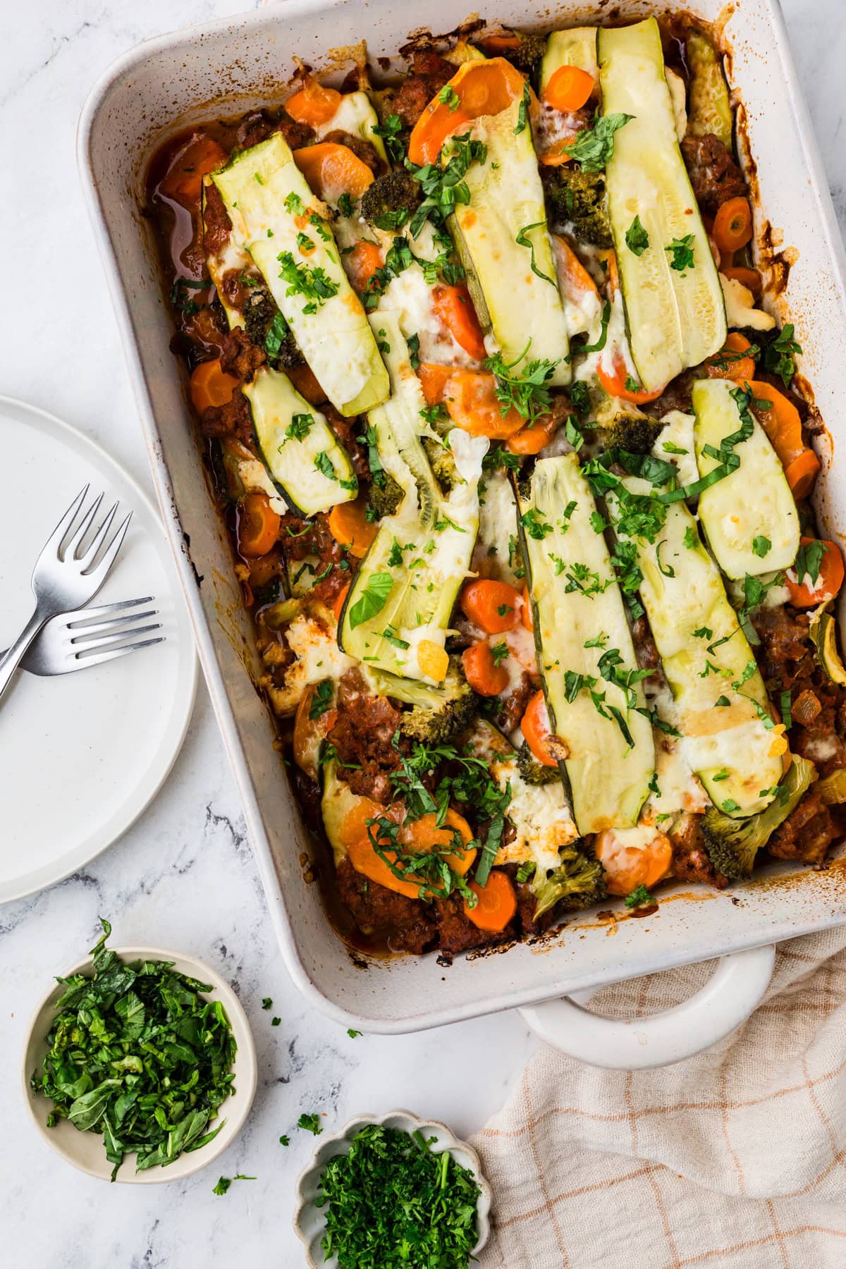 An easy zucchini lasagna stars in a rectangular dish, adorned with sliced zucchini, carrots, broccoli, and melted cheese. Fresh herbs garnish the top. Beside it on the marble surface are a small bowl of chopped herbs and an empty plate with forks.