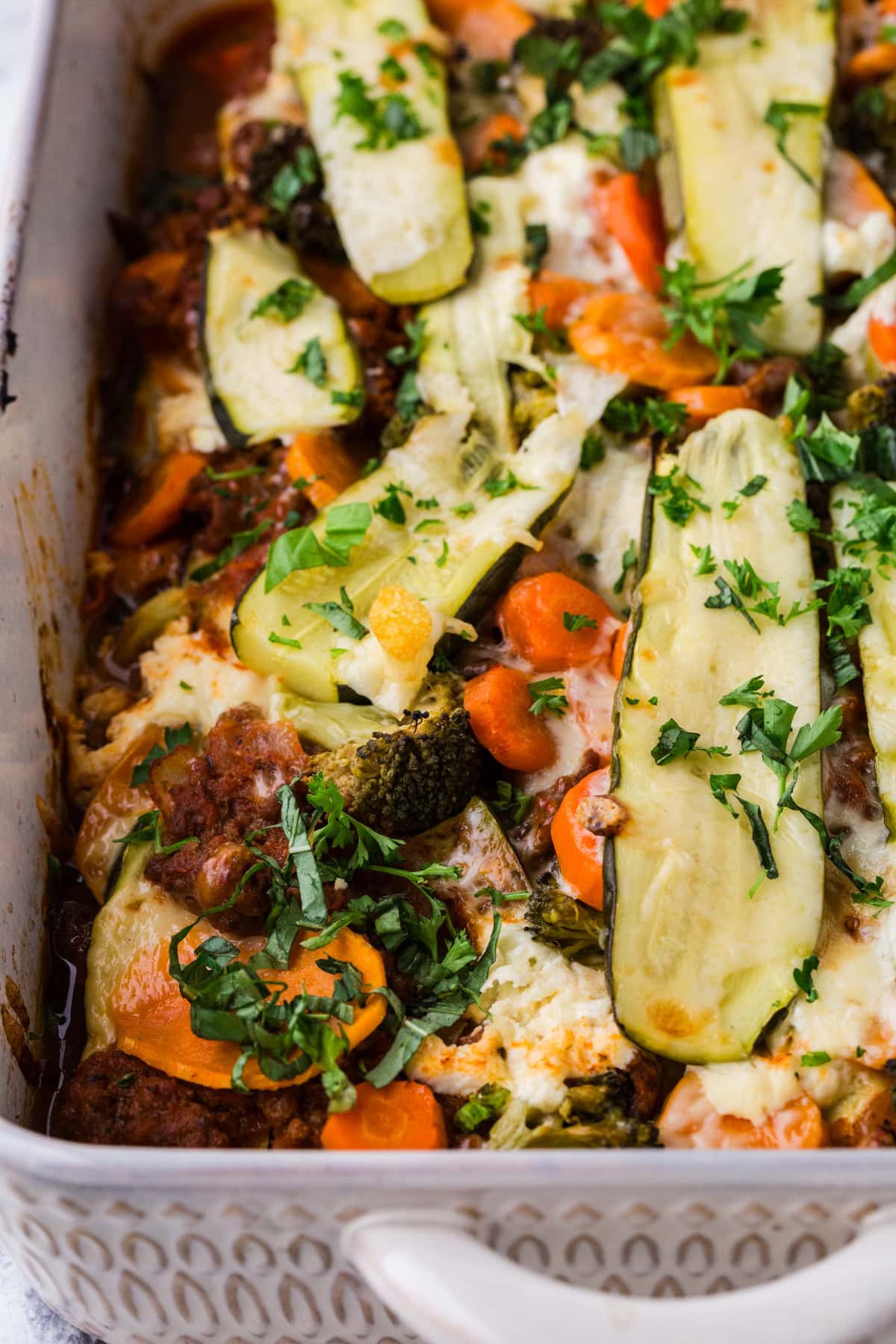 A delightful take on easy zucchini lasagna, this baked vegetable casserole in a white dish showcases layers of melted cheese, sliced zucchini, carrots, broccoli, and herbs. Its elegantly garnished with fresh parsley for an extra touch of flavor.
