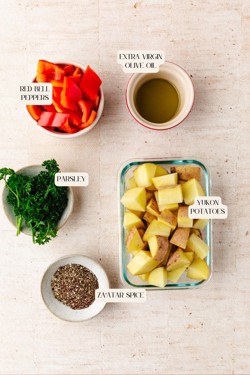 Overhead view of recipe ingredients on a light surface: sliced red bell peppers in a bowl, extra virgin olive oil in a small cup, a bunch of parsley, potatoes in a dish, and spice blend in a small bowl.