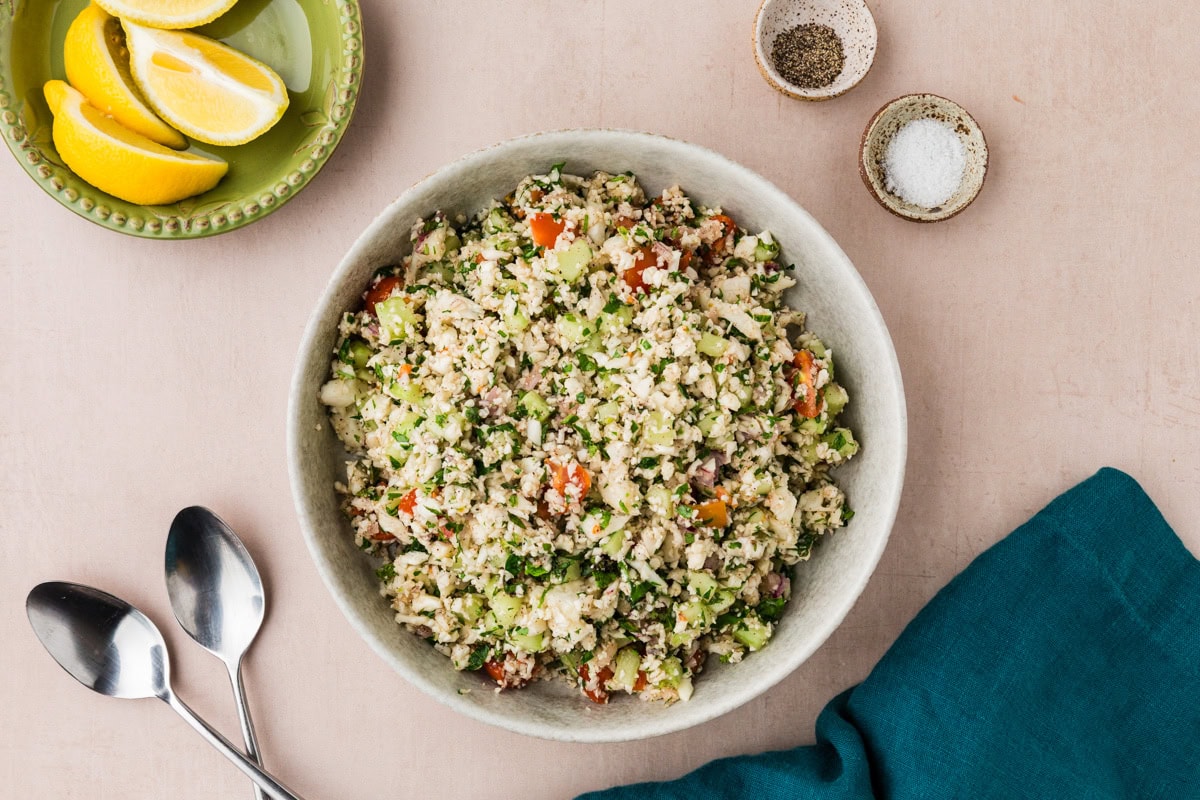 A bowl of fresh Cauliflower Tabbouleh salad with chopped vegetables and herbs sits on a light surface. Nearby are lemon wedges on a green plate, two spoons, plus small bowls of salt and pepper beside a dark teal napkin.