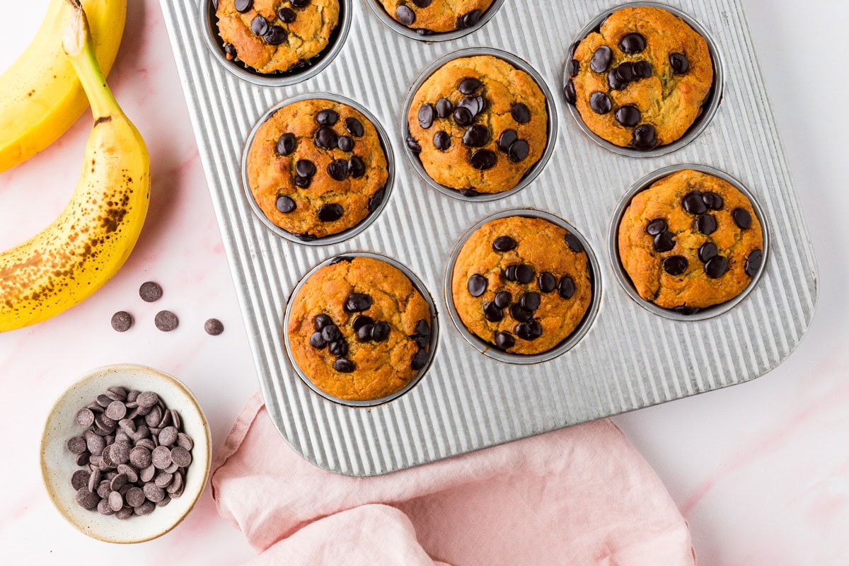 A muffin tray filled with gluten free banana chocolate chip muffins, surrounded by ripe bananas, a bowl of chocolate chips, and a pink cloth on a light surface.