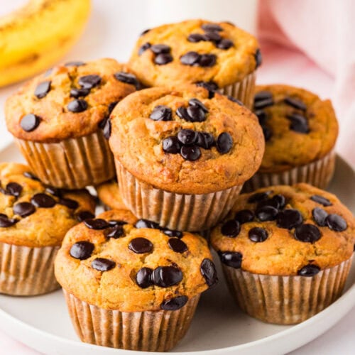 A plate of gluten free banana chocolate chip muffins is stacked on a white plate. A ripe banana and a glass bottle of milk are in the background, with a soft pink cloth partially visible.