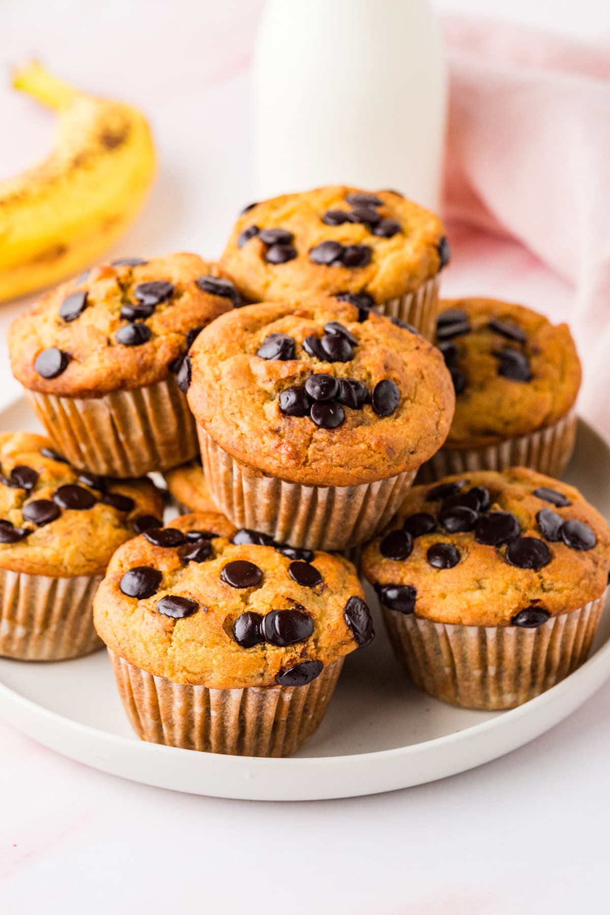A plate of gluten free banana chocolate chip muffins is stacked on a white plate. A ripe banana and a glass bottle of milk are in the background, with a soft pink cloth partially visible.
