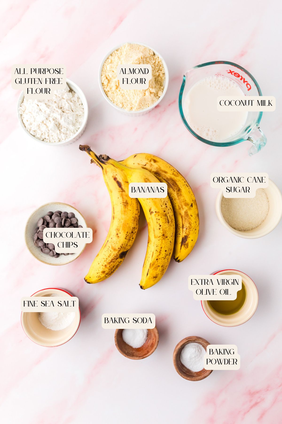 An overhead view of labeled baking ingredients on a pink surface, ready for making gluten free banana chocolate chip muffins, including bananas, gluten free flour, almond flour, coconut milk, organic cane sugar, and chocolate chips.