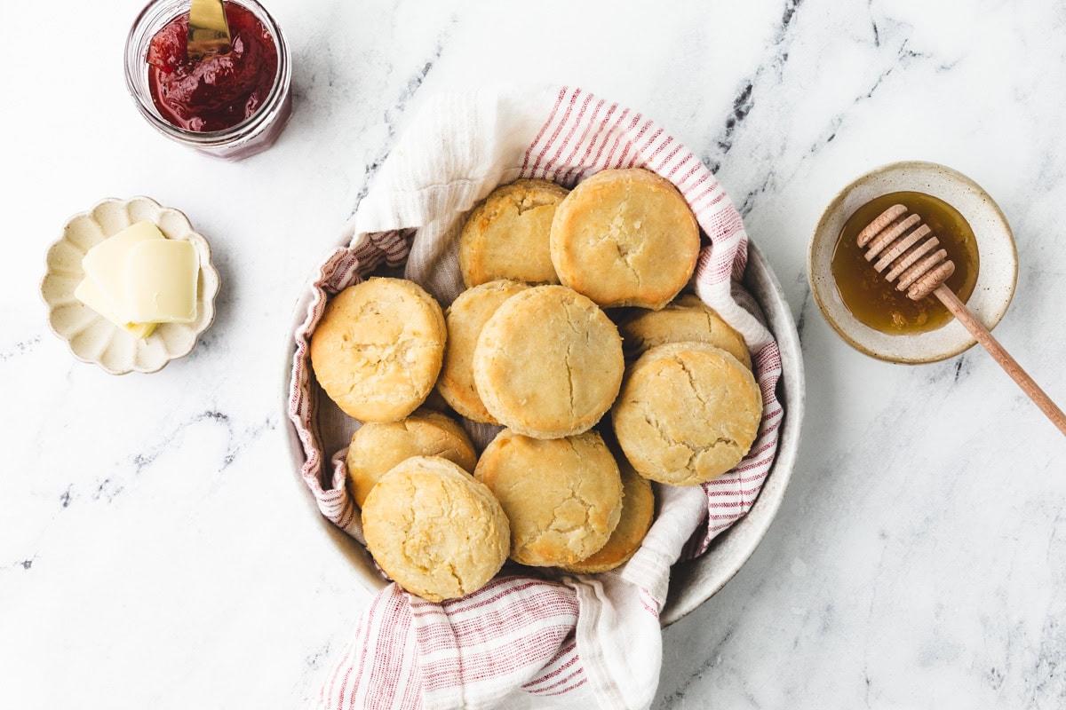 A bowl of freshly baked, gluten-free buttermilk biscuits rests on a cloth atop a marble surface. Nearby, a plate with butter pats, a jar of jam, and a bowl with honey and a wooden dipper await to complement the delightful treat.