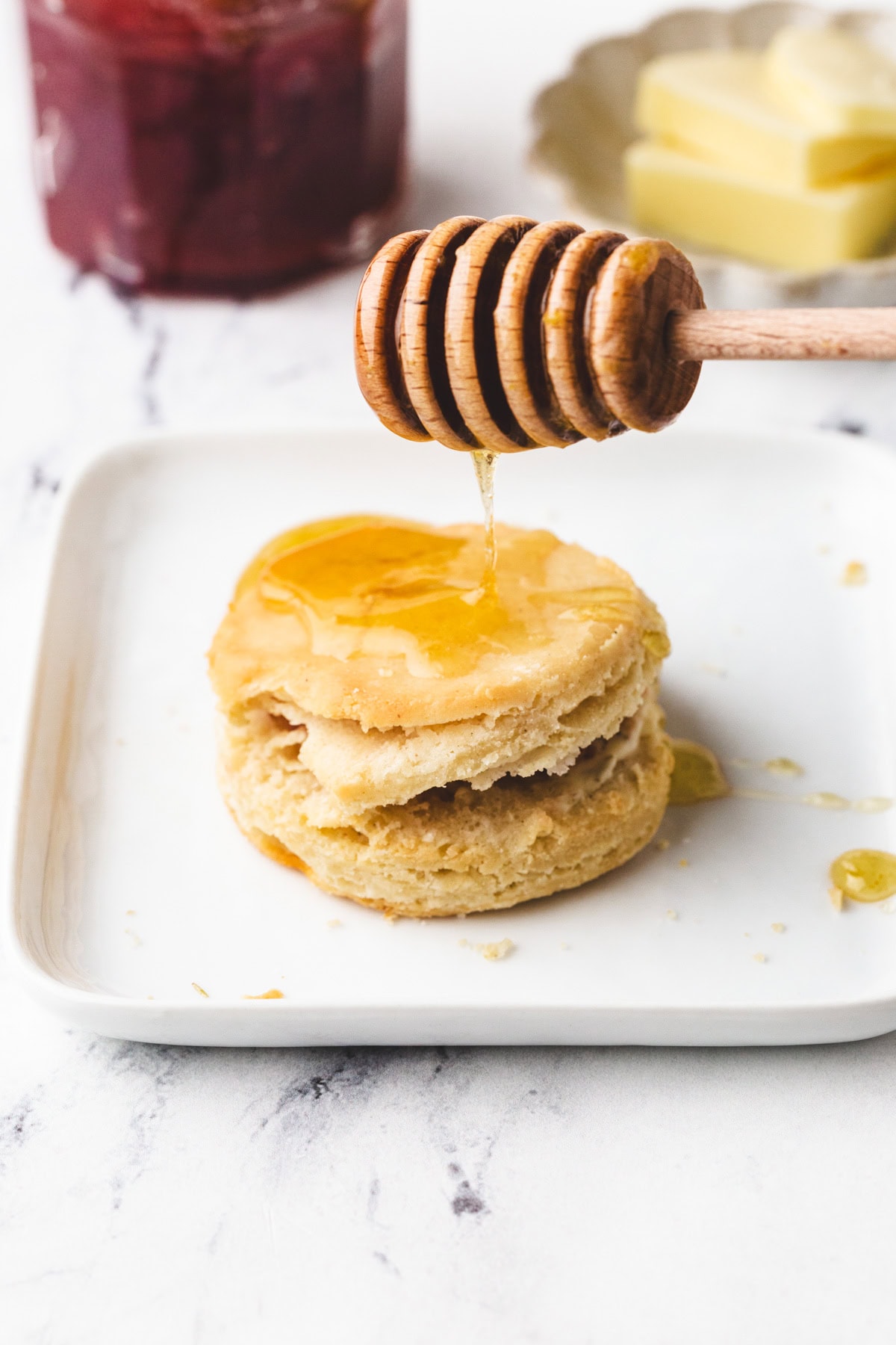 A honey dipper drizzles honey onto a gluten-free buttermilk biscuit stacked on a white plate. In the background, theres a jar of red jam and a small dish with butter, all set on a marble surface.
