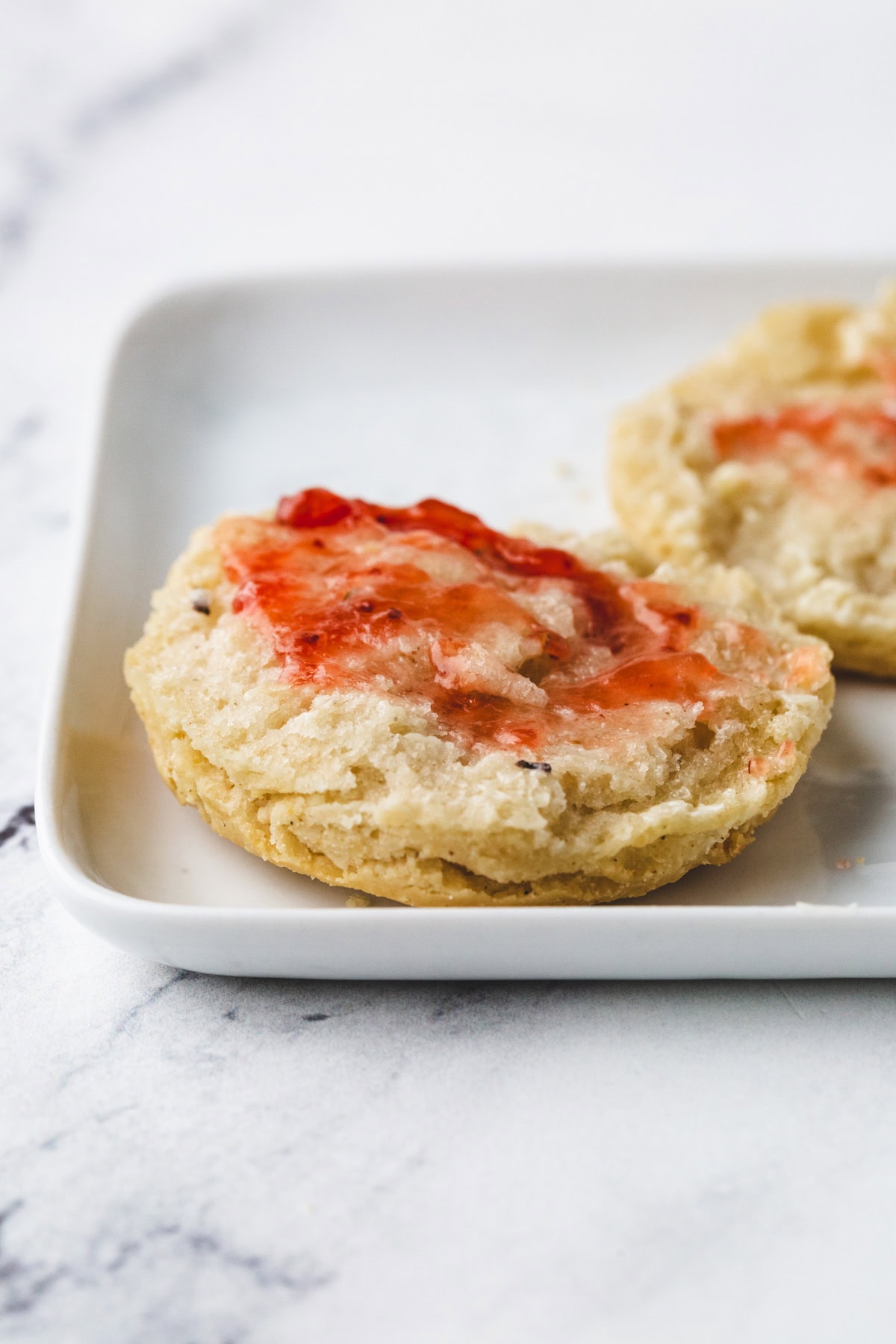 A close-up of a gluten-free biscuit topped with melted butter and strawberry jam on a square white plate. The biscuit has a flaky texture and light golden color, thanks to its buttermilk batter. The background features a pristine white marble surface.