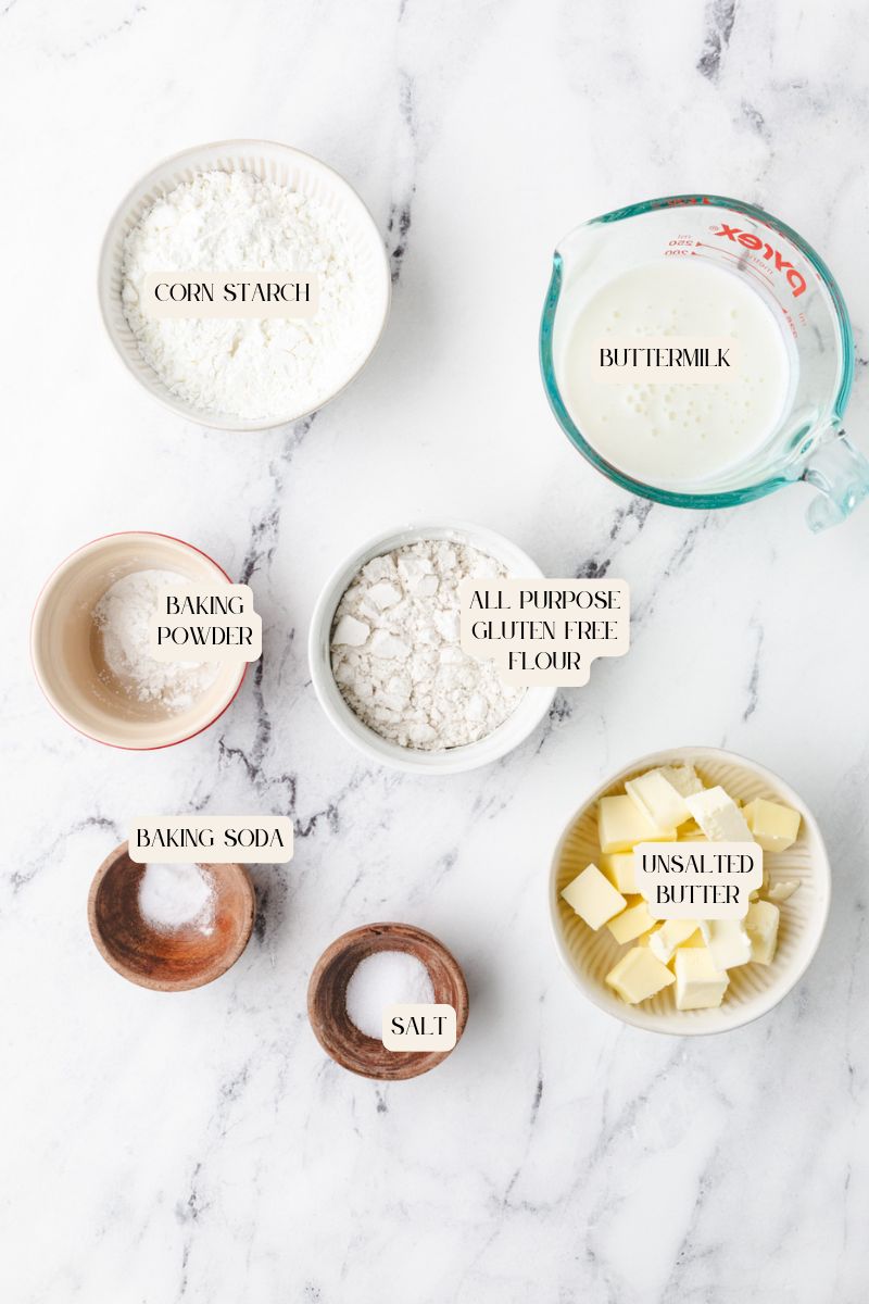 A top-view of labeled baking ingredients for gluten-free buttermilk biscuits on a marble surface: corn starch, baking powder, baking soda, salt, all-purpose gluten-free flour, unsalted butter, and a measuring cup of buttermilk.