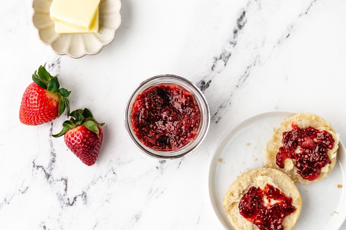 A top view of a breakfast setting on a marble surface. A jar of strawberry jam is surrounded by fresh strawberries, pats of butter on a dish, and a plate with two halves of an English muffin spread with strawberry jam.