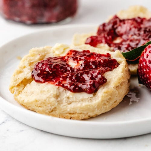 A close-up of a scone halved on a white plate, topped with butter and strawberry jam. A fresh strawberry rests beside it. A jar of jam is blurred in the background.