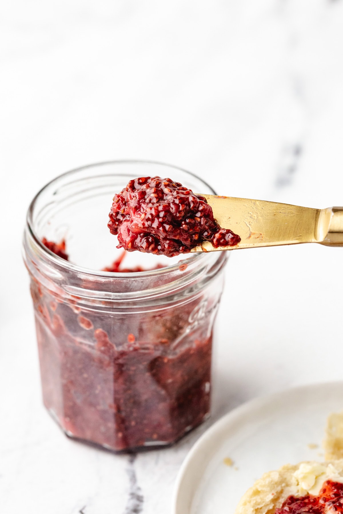 A jar of Strawberry Preserves Without Sugar is open on a white surface. A gold knife holds a dollop of the jam above the jar. A partially visible plate with bread is in the lower right corner.