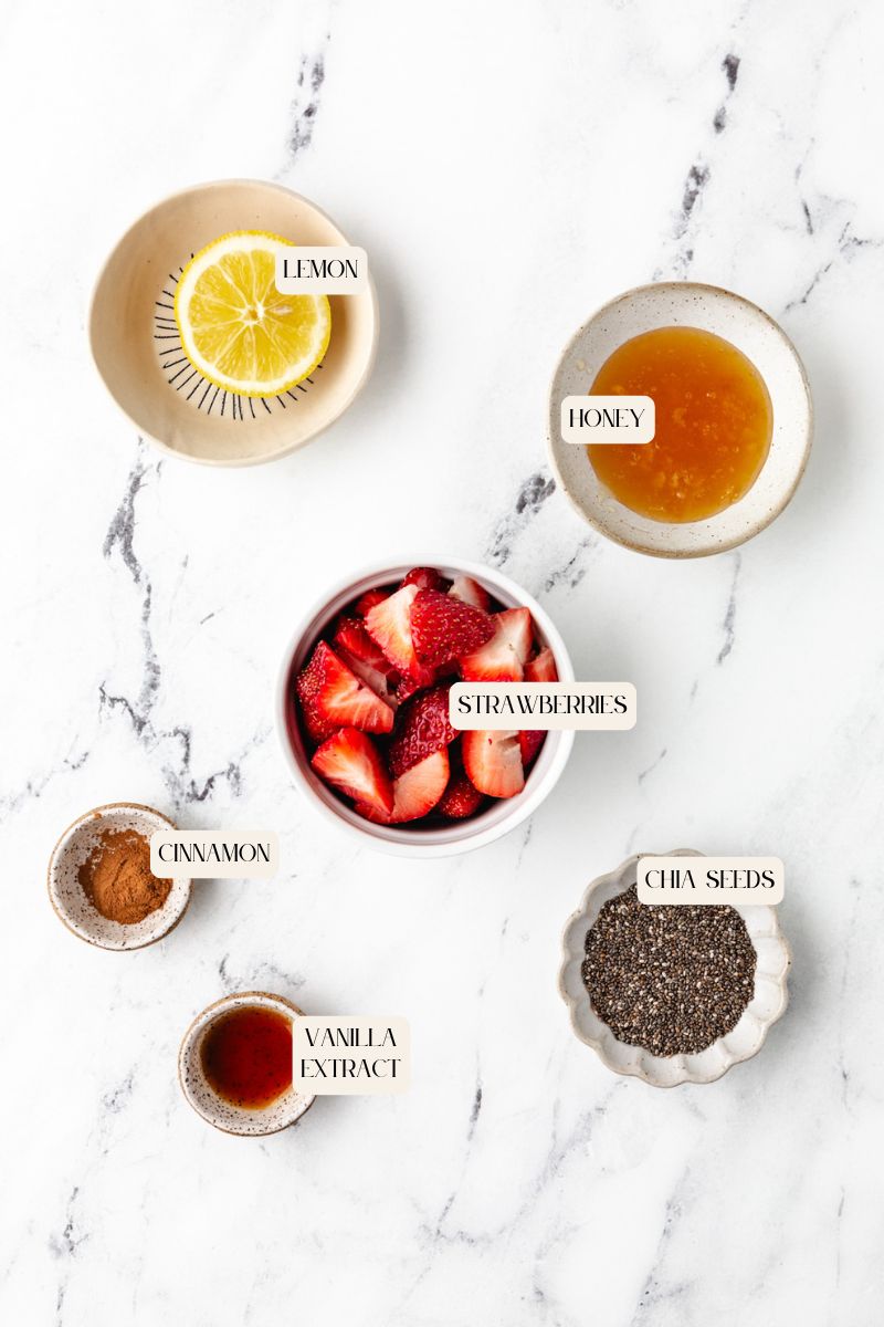 Top view of ingredients on a marble surface: a lemon slice in a bowl, honey in a bowl, sliced strawberries hinting at sugar-free strawberry preserves, chia seeds in a dish, vanilla extract in a small dish, and cinnamon powder in a bowl. Labels identify each ingredient.