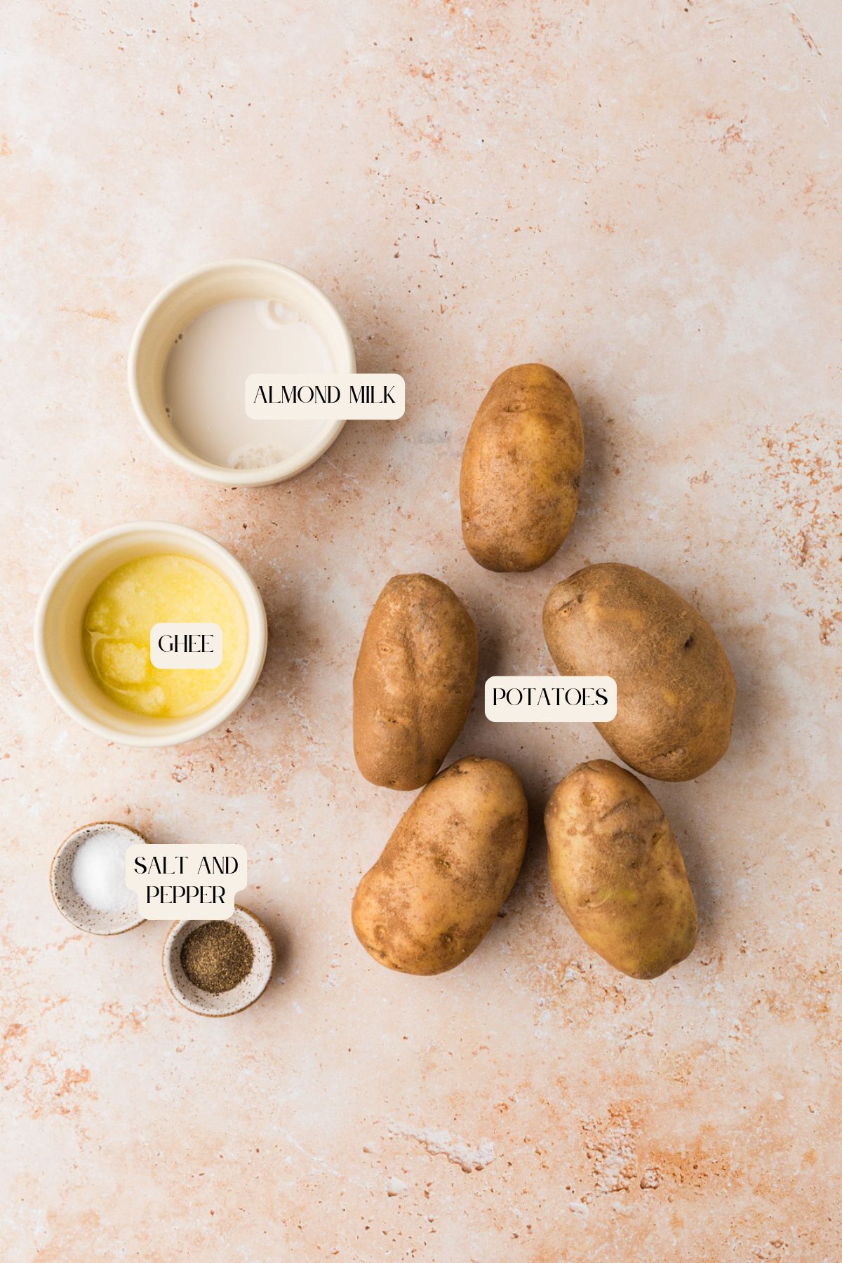 Five russet potatoes, a small bowl of almond milk, a small bowl of melted ghee, and two small bowls with salt and pepper are arranged on a light, textured surface. Each item is labeled.