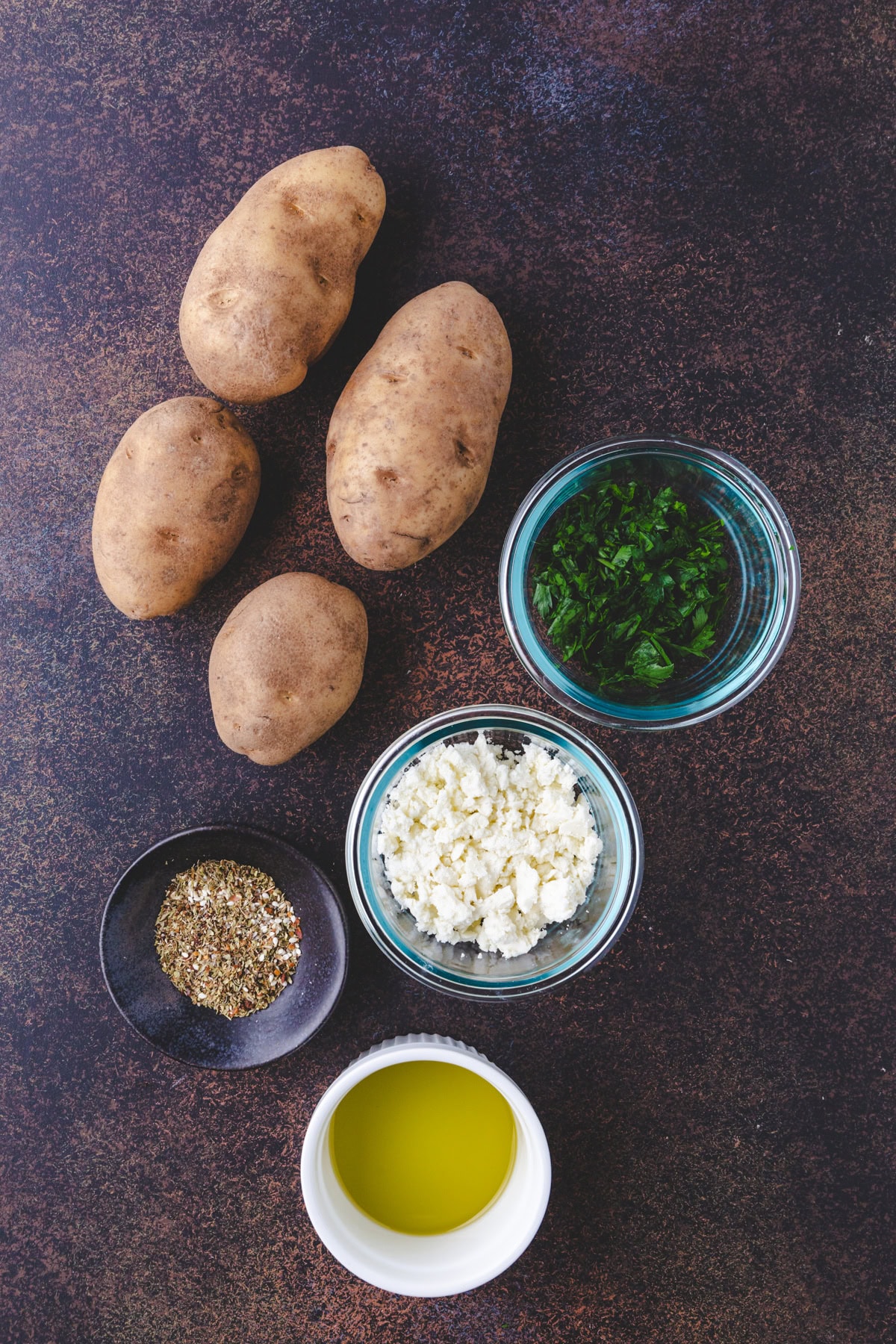 Four raw potatoes, a bowl of chopped herbs, crumbled cheese, oil, and a small dish of mixed spices arranged on a dark countertop—everything you need for delicious zaatar fries.