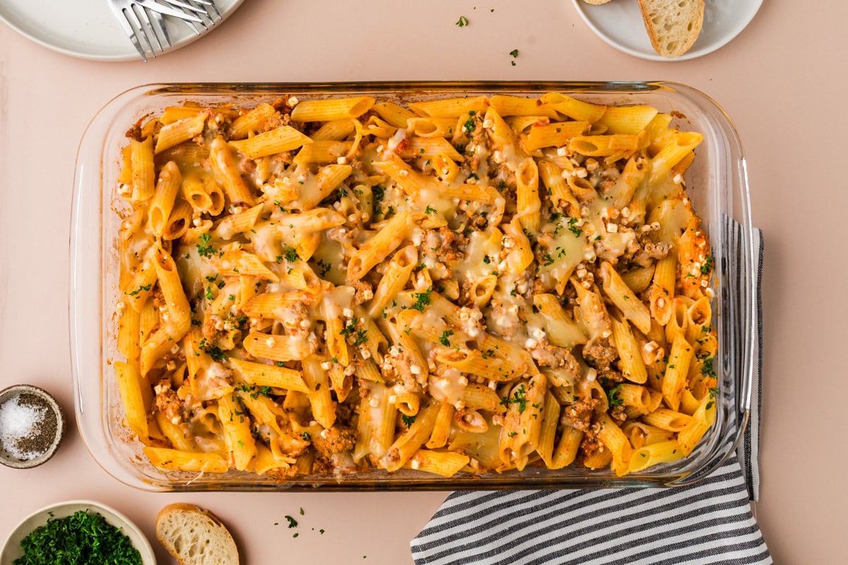 Baked penne pasta with melted cheese and ground meat in a glass dish, just like a 5 ingredient baked ziti, garnished with chopped herbs, next to slices of bread, a plate, and a striped napkin on a beige surface.