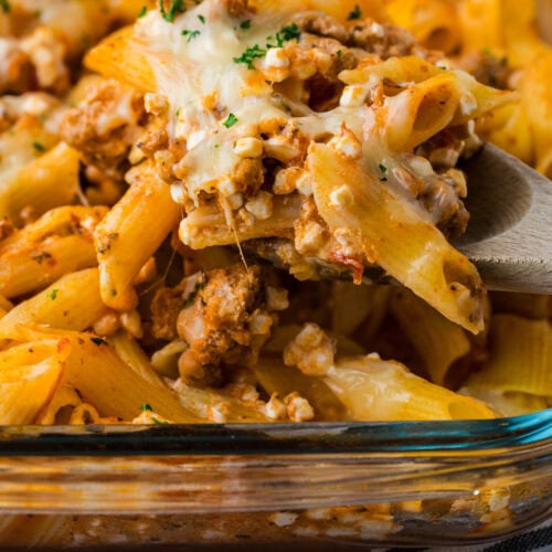 A close-up of a 5 ingredient baked ziti casserole with penne, ground meat, tomato sauce, and melted cheese, being served with a wooden spoon from a glass baking dish.