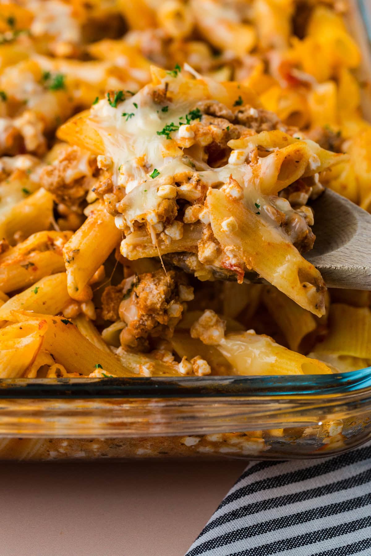 A close-up of a 5 ingredient baked ziti casserole with penne, ground meat, tomato sauce, and melted cheese, being served with a wooden spoon from a glass baking dish.