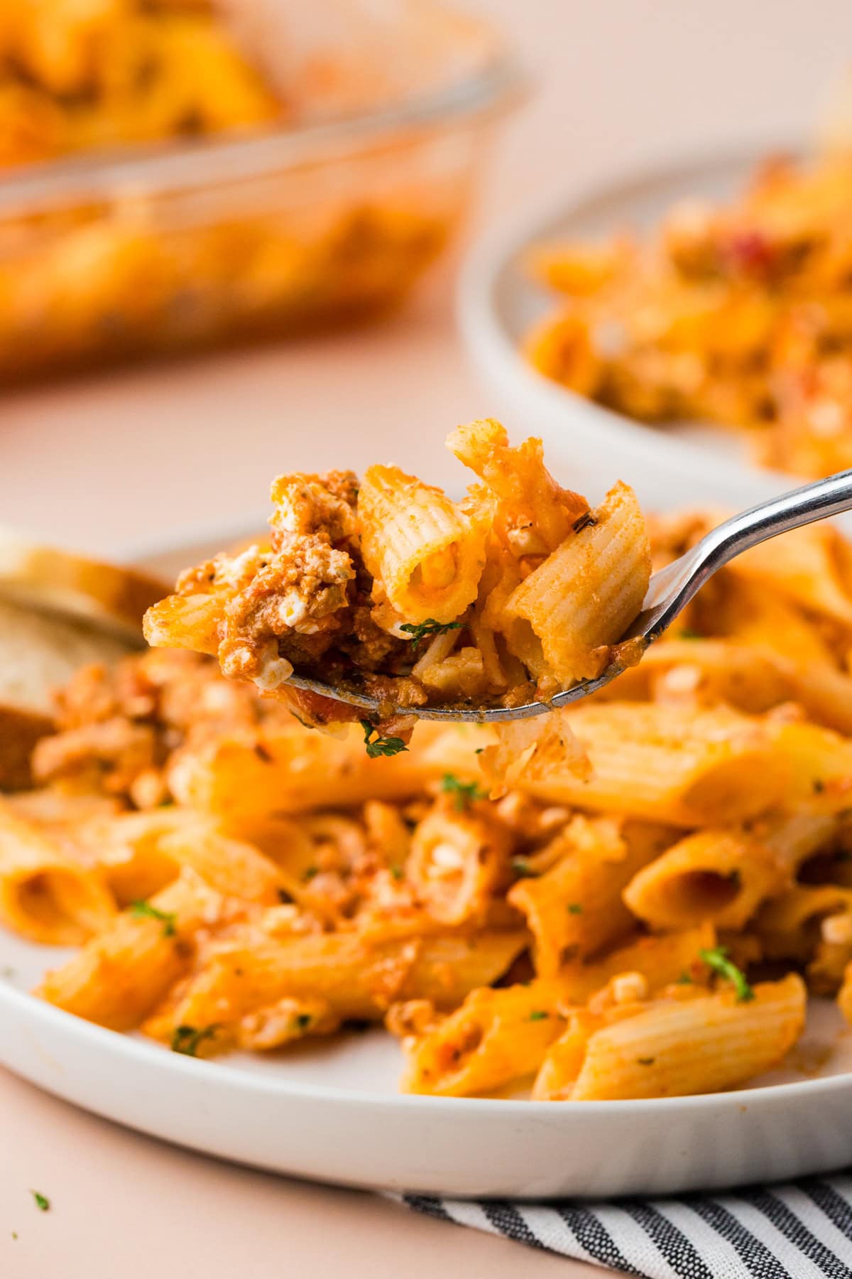 A close-up of a fork holding a bite of cheesy 5 ingredient baked ziti with tomato sauce, with more pasta served on a white plate in the background.