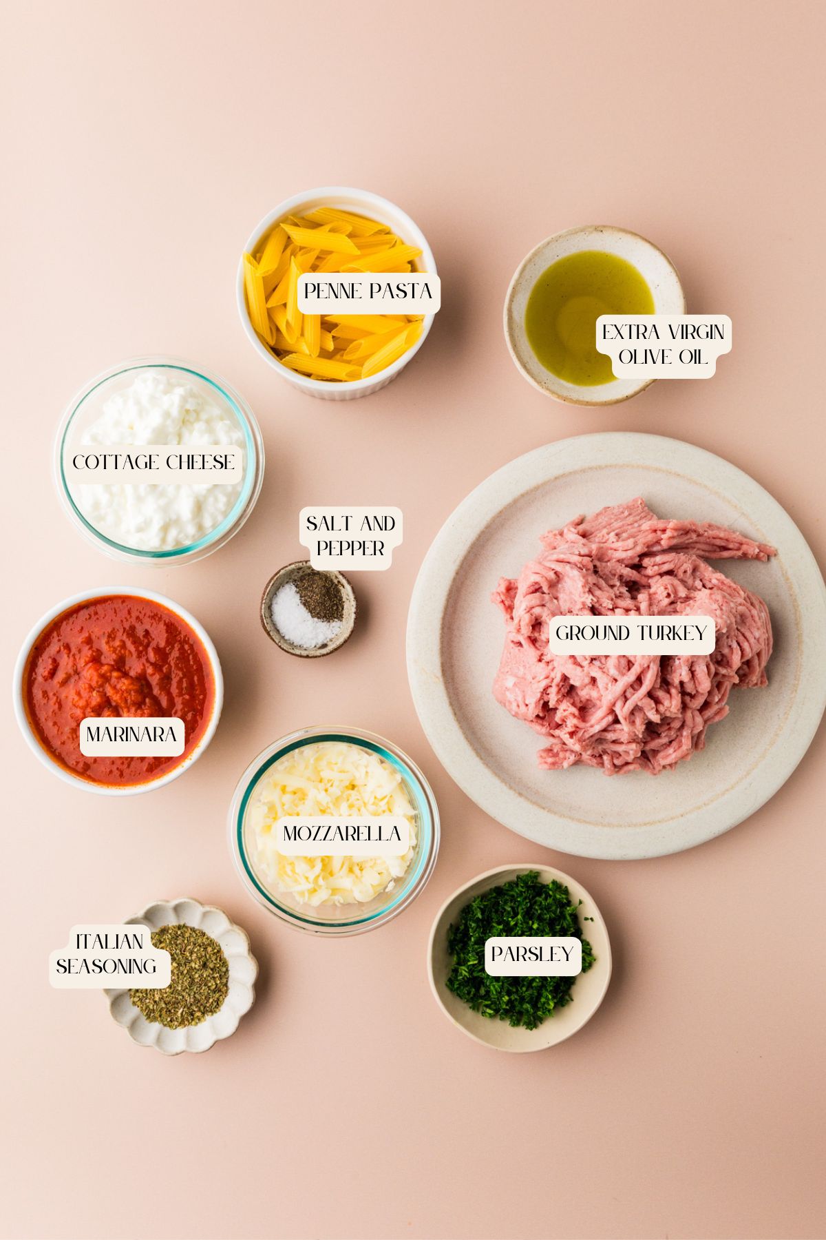 Ingredients for 5 ingredient baked ziti are displayed on a light pink surface, including penne pasta, cottage cheese, marinara sauce, mozzarella, ground turkey, and more. Each item is clearly labeled.