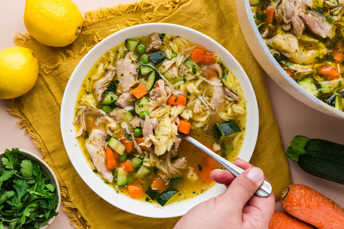A hand holds a spoon over a bowl of chicken soup with orzo, carrots, zucchini, and herbs, set on a yellow cloth. Lemons, cilantro, zucchini, and carrots are nearby, along with a pot of soup.