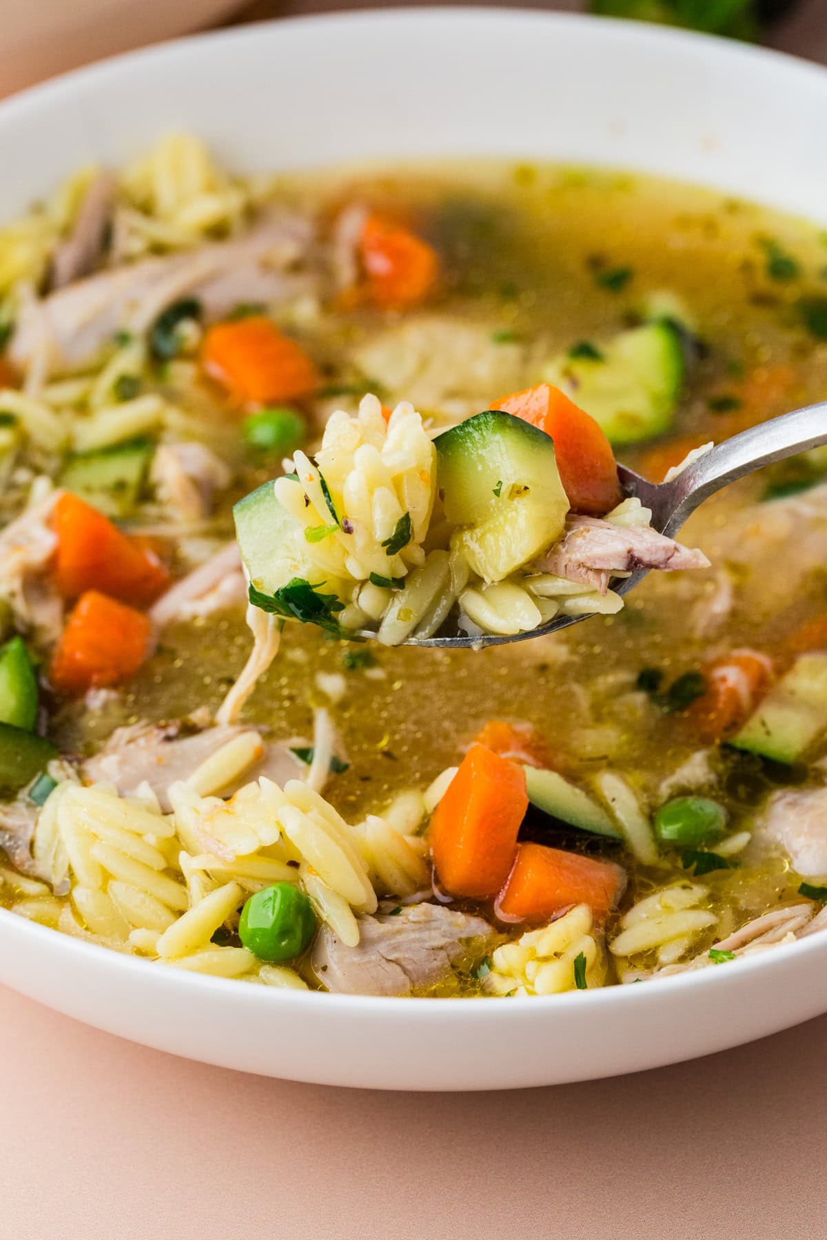 A close-up of a spoonful of vegetable and orzo soup with carrots, zucchini, peas, and shredded meat, held above a white bowl filled with the same soup.