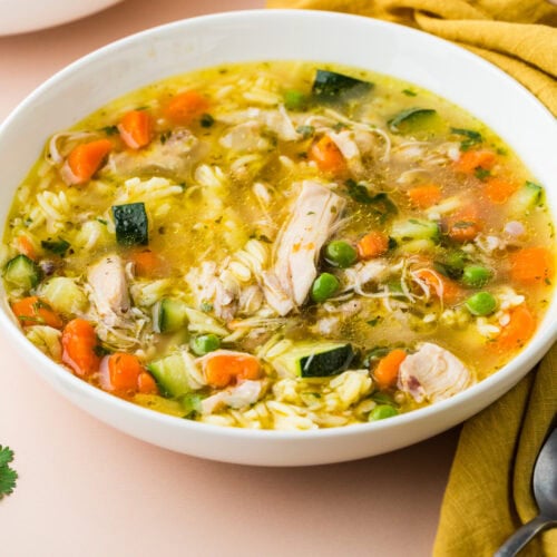 A white bowl of chicken and vegetable soup with carrots, peas, zucchini, rice, and herbs, placed on a light surface next to a mustard yellow napkin and a silver spoon.