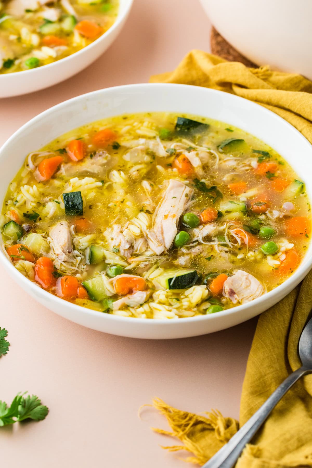 A white bowl of chicken and vegetable soup with carrots, peas, zucchini, rice, and herbs, placed on a light surface next to a mustard yellow napkin and a silver spoon.