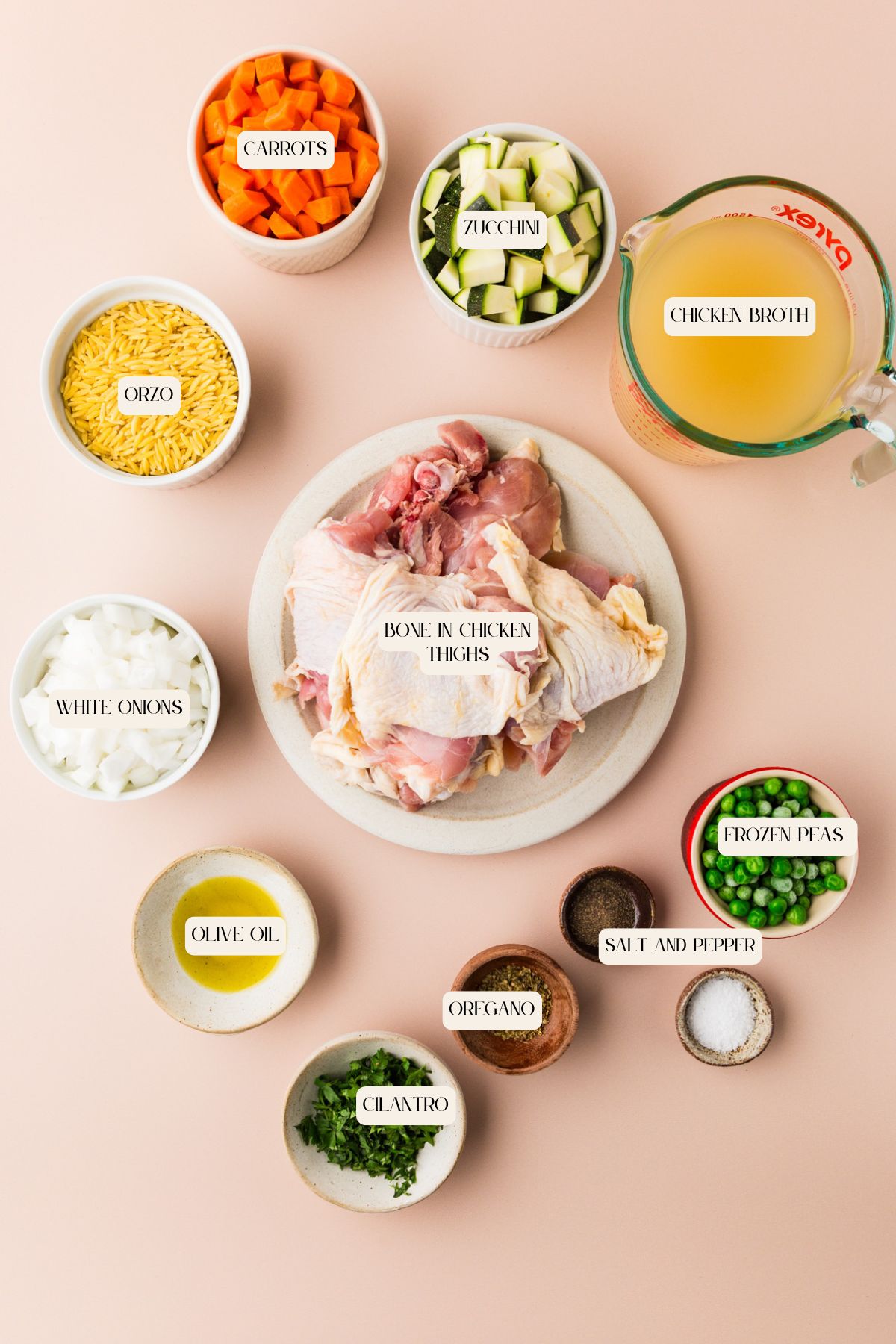 A flat lay of soup ingredients: a plate of raw bone-in chicken thighs, bowls of chopped carrots, zucchini, white onions, orzo, frozen peas, cilantro, oregano, salt and pepper, olive oil, and a measuring cup of chicken broth.