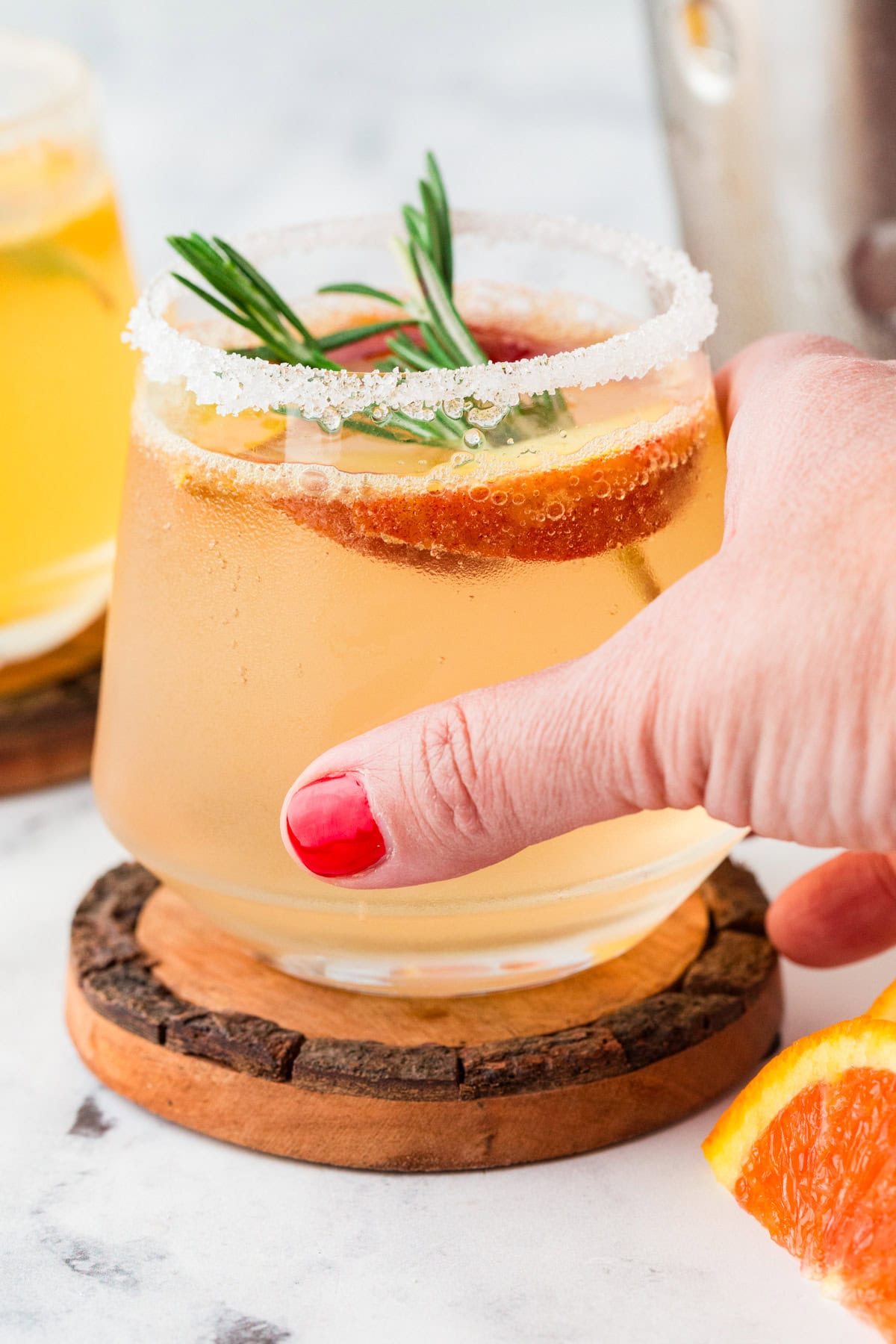 A hand with red nail polish holds a glass of white sangria mocktail, garnished with a slice of orange and a sprig of rosemary. The sparkling drink has a sugared rim and rests on a wooden coaster.